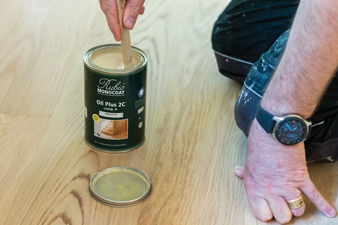 Person using a stirring stick to apply Rubio Monocoat Oil Plus 2C on a wooden floor.